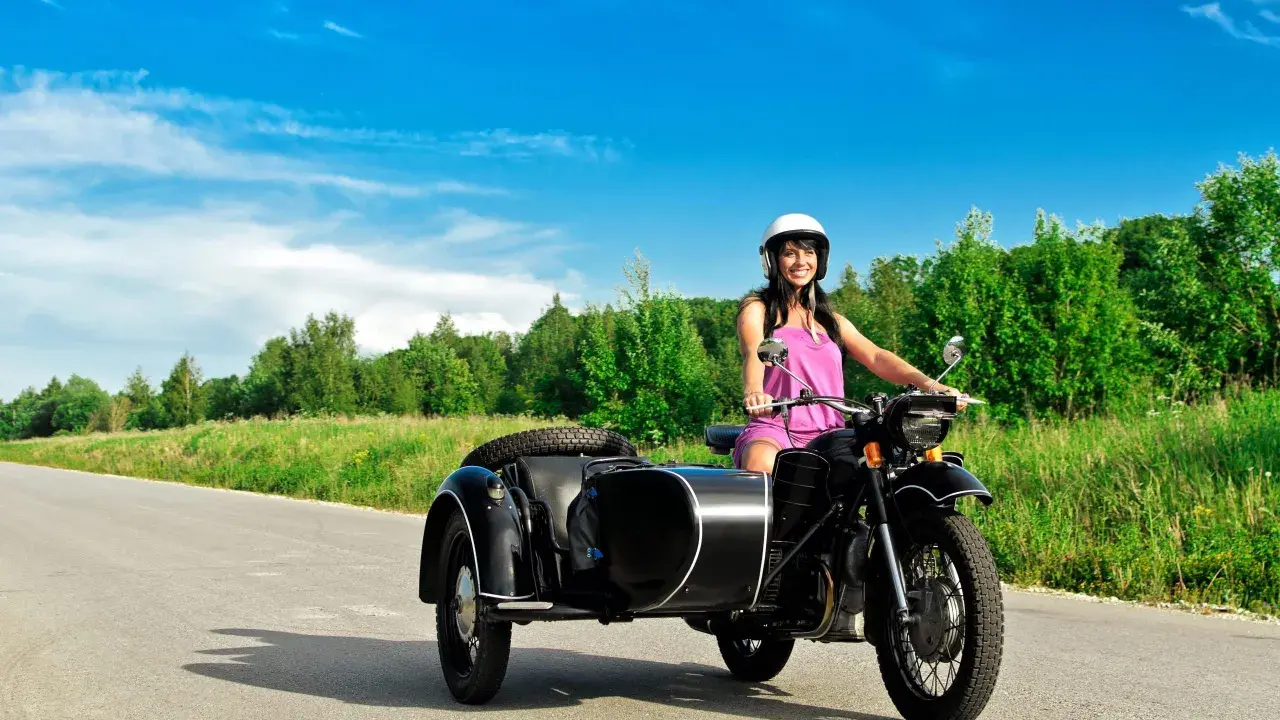 Mujer sonriente con casco blanco conduce una motocicleta negra con sidecar. El permiso A1 autoriza a conducir motocicletas con sidecar.