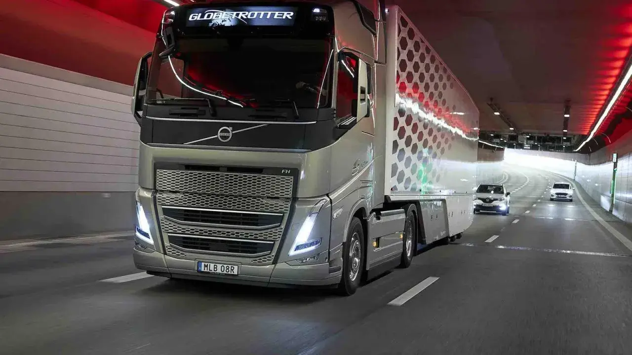 Camión Volvo FH Globetrotter plateado avanza por un túnel con luces rojas. El camión, que parece ser un modelo i-Save, transporta una carga con un diseño hexagonal.