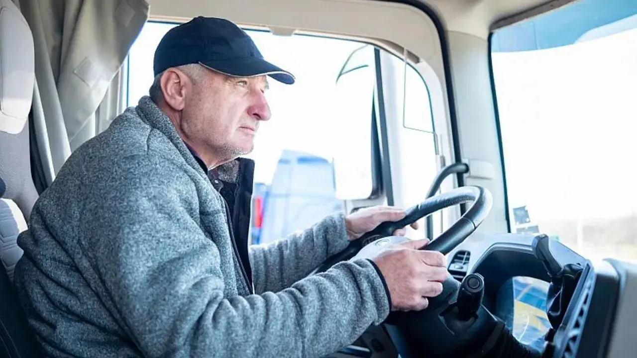 Conductor de camión con gorra y chaqueta gris, con las manos en el volante. Podría ser parte de un listado de cese de actividad de transportistas.