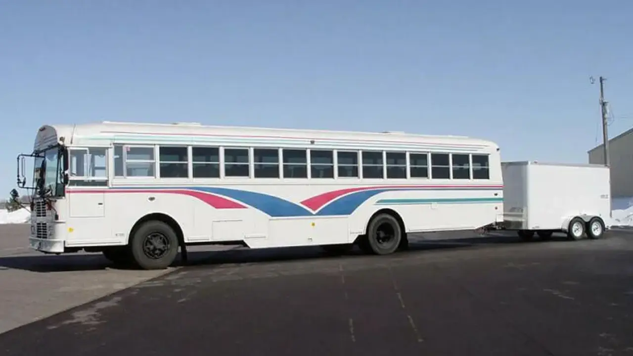 Autobús blanco con franjas azules y rosas, remolcando un tráiler. Ideal para un viaje largo con el carnet D+E.