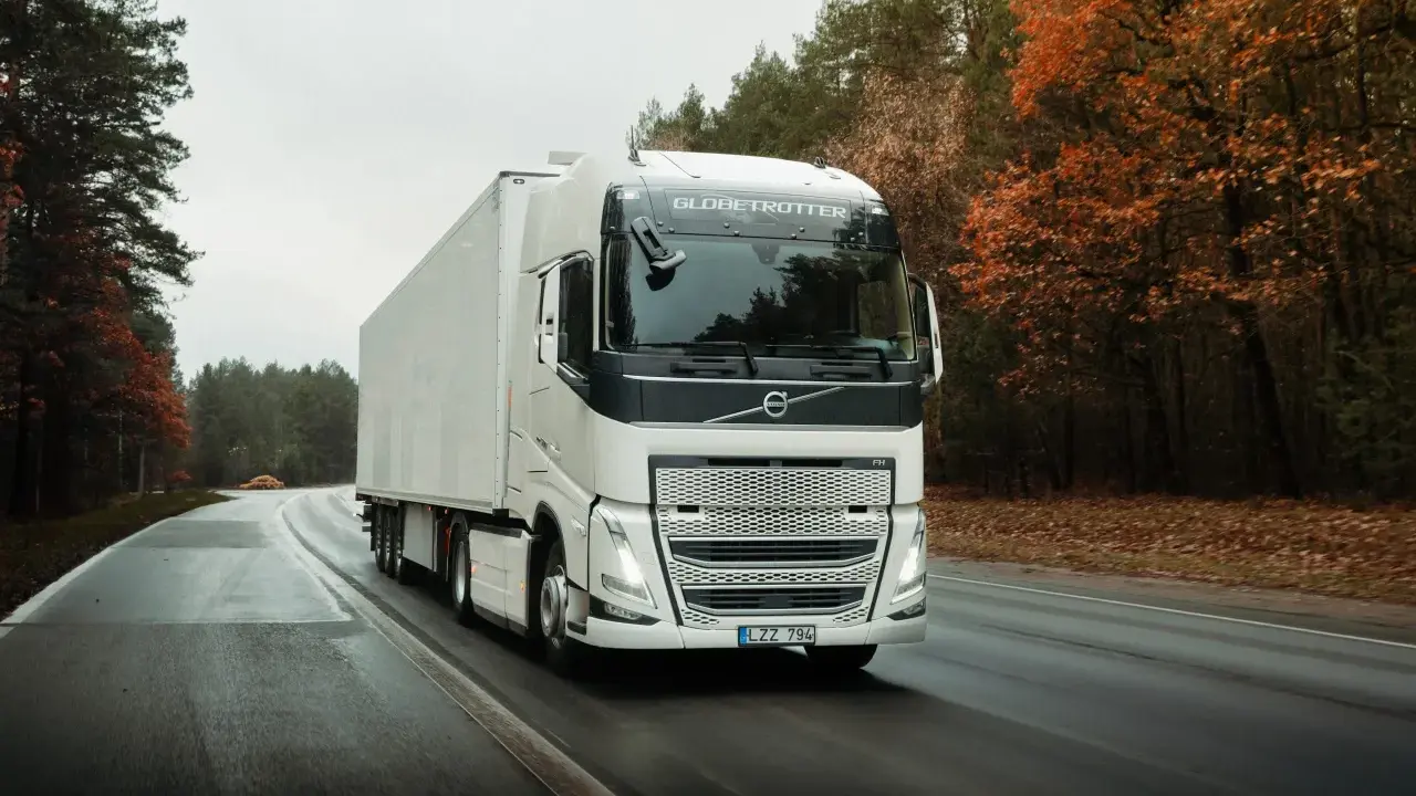 Un camión Volvo FH blanco con remolque blanco circula por una carretera mojada rodeada de árboles otoñales.