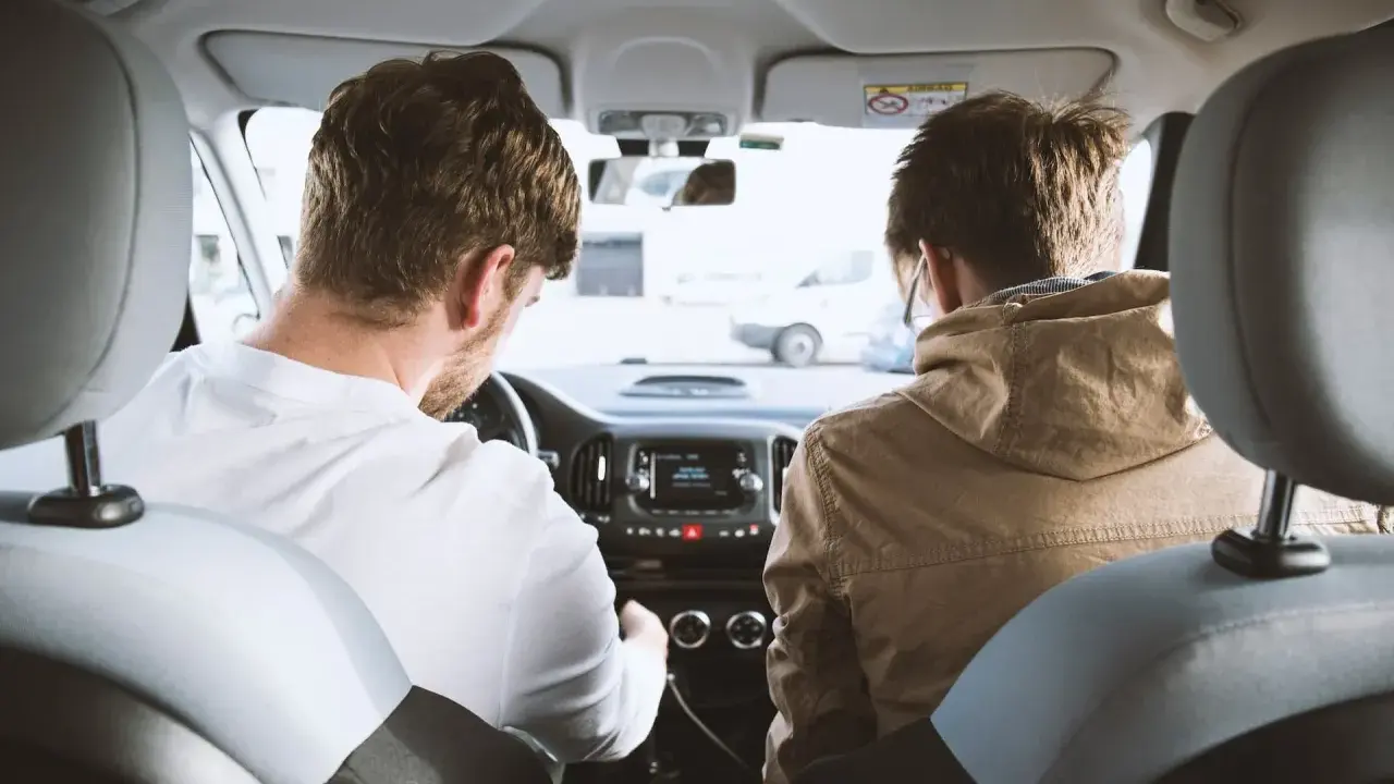 Dos hombres en un coche, uno conduce y el otro mira. Podrían estar discutiendo los tipos de faltas que se cometen al volante.