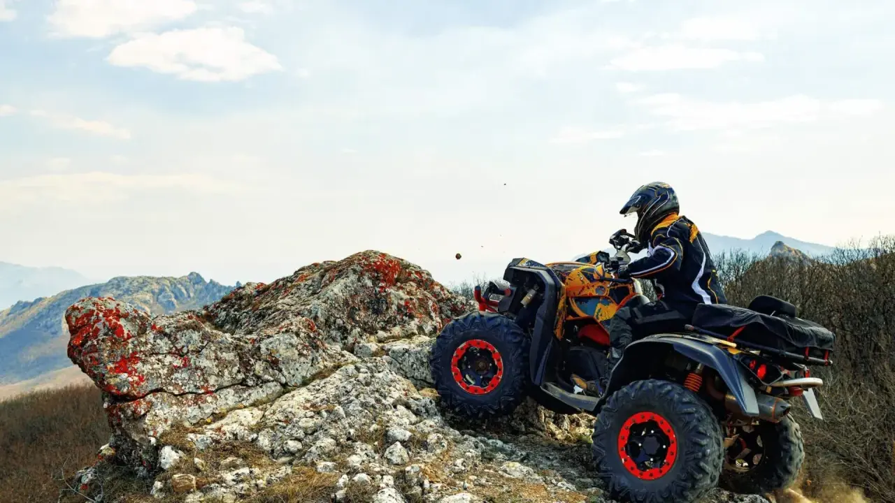 Un piloto con casco y chaqueta amarilla y negra conduce un quad ATV por un terreno rocoso y montañoso. El permiso para esta aventura es la emoción.