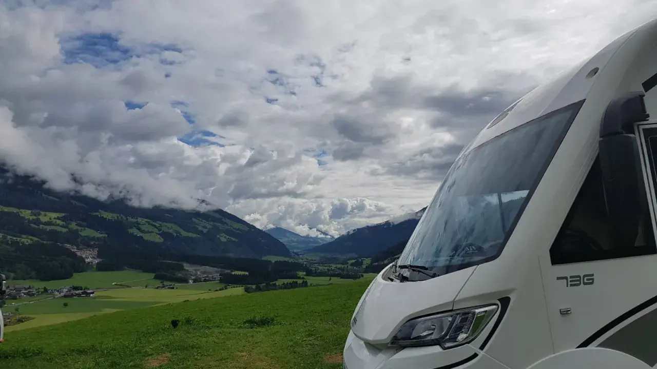 Autocaravana de más de 3500 kg aparcada en un prado verde con vistas a un valle alpino y nubes dramáticas.