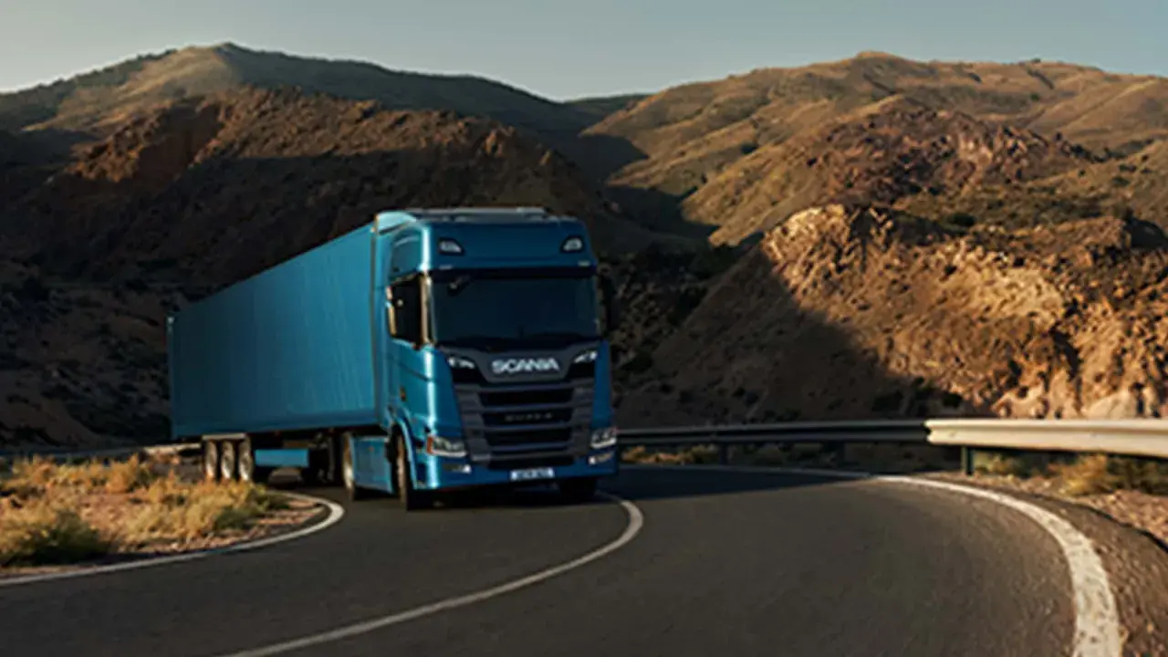 Camión Scania azul recorre una carretera sinuosa entre montañas áridas, evocando la fuerza y el espíritu de aventura de Suecia.