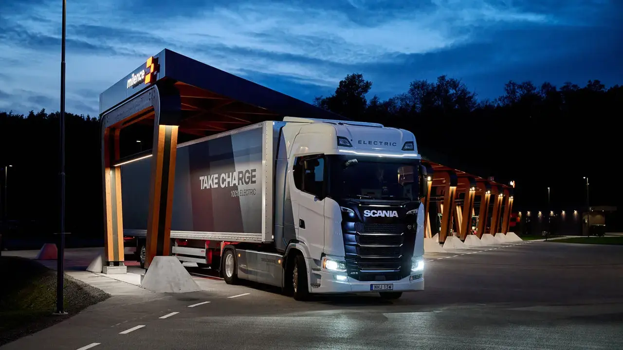 Camión Scania eléctrico en una estación de carga nocturna. El remolque dice "TAKE CHARGE 100% ELECTRIC".