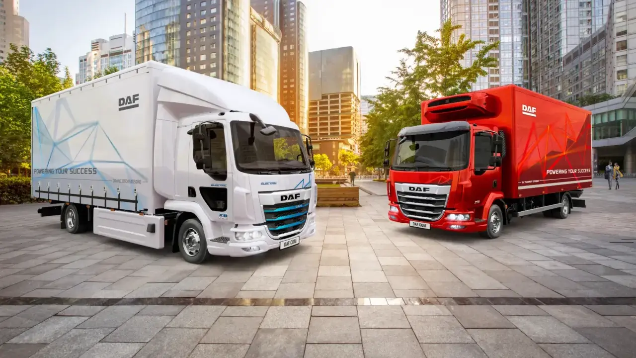 Dos camiones DAF, uno blanco eléctrico y otro rojo, en una ciudad moderna. El DAF XB blanco muestra "POWERING YOUR SUCCESS".