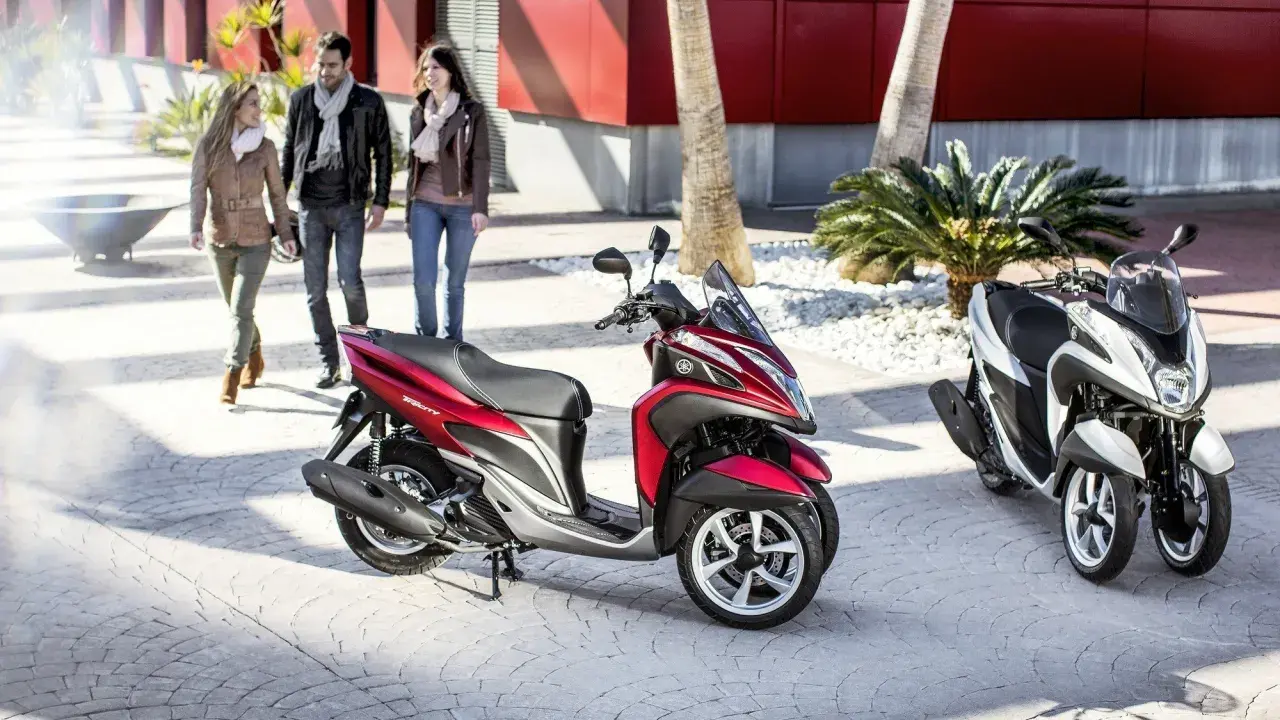 Dos motocicletas Yamaha Tricity, una roja y otra blanca, listas para circular. Tres personas pasean al fondo.