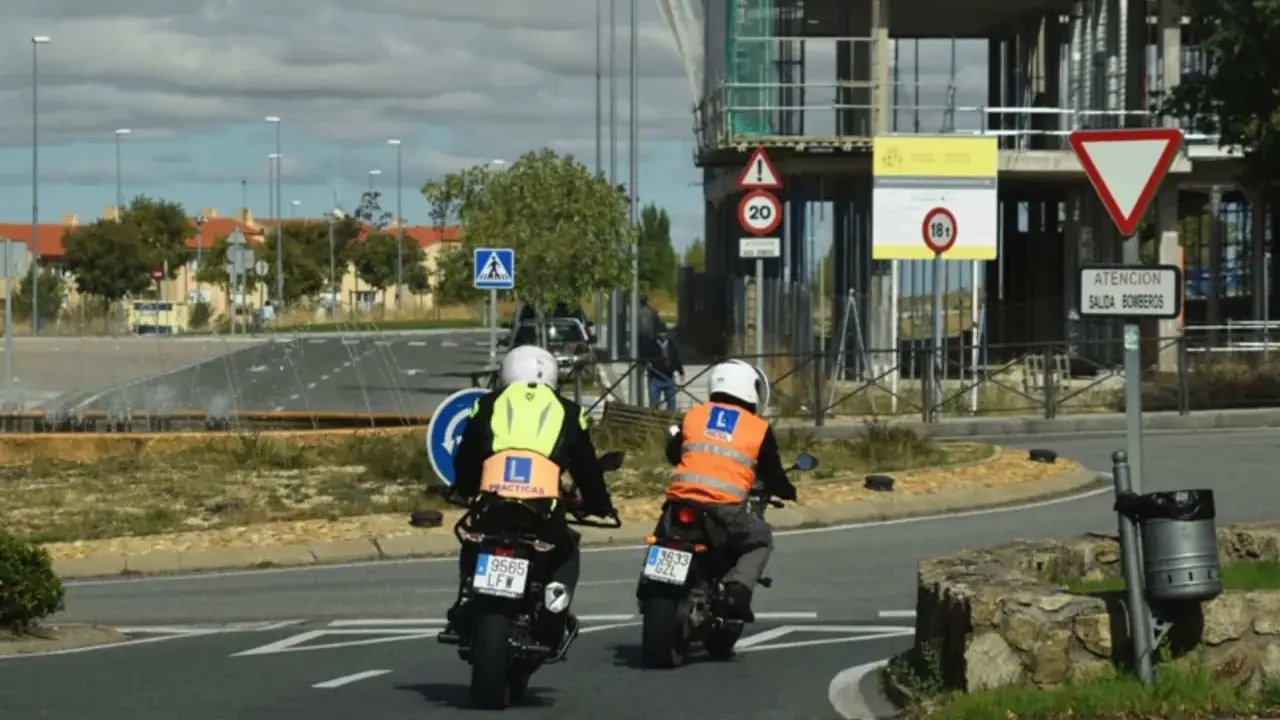 Dos motoristas con chalecos de prácticas, uno amarillo y otro naranja, circulan en moto. Uno de ellos lleva un cartel de "carnet moto 300".