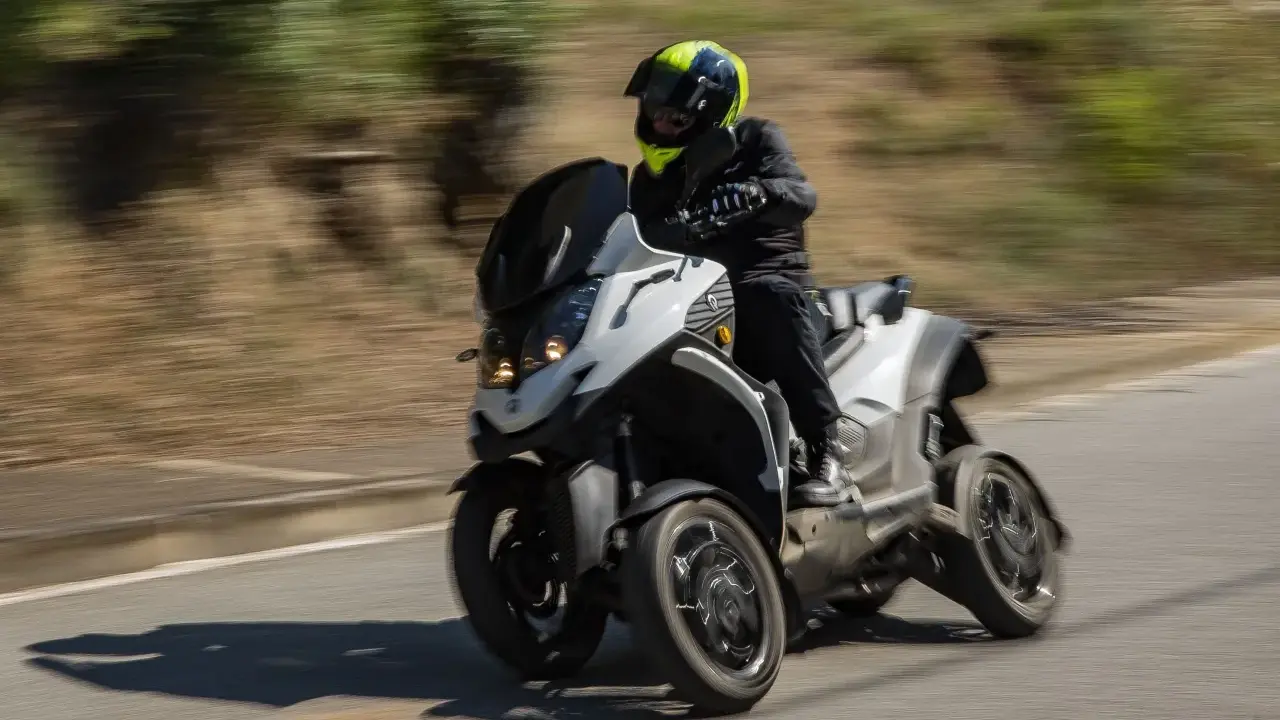 Un motorista con casco amarillo y negro conduce una moto de tres ruedas por una carretera. La moto ciudad se mueve rápido, creando un efecto de desenfoque.