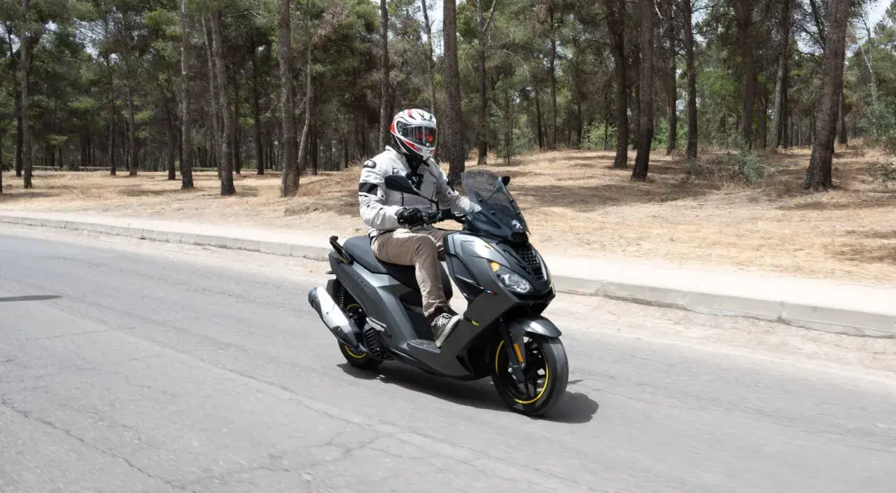 Hombre en casco y chaqueta conduce un triciclo de motor gris por una carretera rodeada de pinos.