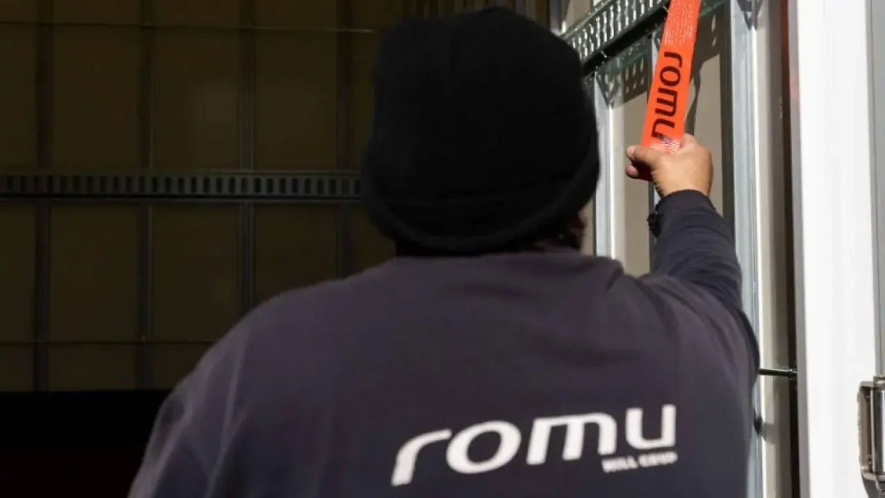 Persona con gorro negro ajusta una cinta naranja con el logo "romu", evitando una multa por exceso de peso.