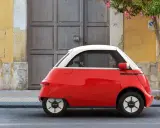 Un triciclo y cuadriciclo de motor rojo y blanco, un vehículo eléctrico compacto, estacionado frente a una puerta de madera y una pared amarilla.