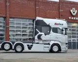 Camión Scania Longline plateado con diseño negro y rojo, aparcado frente a un edificio de ladrillo con puertas de garaje.