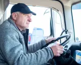 Conductor de camión con gorra y chaqueta gris, con las manos en el volante. Podría ser parte de un listado de cese de actividad de transportistas.