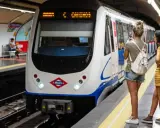 En la estación de metro, un tren blanco espera. Varias personas observan, una podría recibir una multa por usar tarjeta de transporte de otra persona.