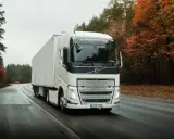 Un camión Volvo FH blanco con remolque blanco circula por una carretera mojada rodeada de árboles otoñales.