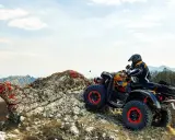 Un piloto con casco y chaqueta amarilla y negra conduce un quad ATV por un terreno rocoso y montañoso. El permiso para esta aventura es la emoción.