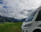 Autocaravana de más de 3500 kg aparcada en un prado verde con vistas a un valle alpino y nubes dramáticas.