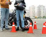 Persona se prepara para el circuito examen moto a1. Un instructor revisa una lista junto a una moto y conos naranjas.