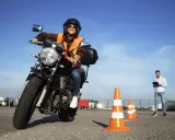 Mujer sonriente practica en circuito cerrado a1, con conos naranjas y un instructor al fondo.