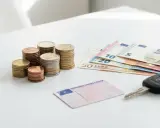 Monedas, billetes de euro, un carnet de precio y llaves de coche sobre una mesa blanca.