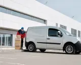 Un repartidor con mascarilla apila cajas junto a una furgoneta plateada frente a un edificio moderno.
