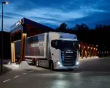 Camión Scania eléctrico en una estación de carga nocturna. El remolque dice "TAKE CHARGE 100% ELECTRIC".
