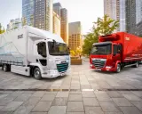 Dos camiones DAF, uno blanco eléctrico y otro rojo, en una ciudad moderna. El DAF XB blanco muestra "POWERING YOUR SUCCESS".