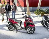 Dos motocicletas Yamaha Tricity, una roja y otra blanca, listas para circular. Tres personas pasean al fondo.