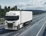Un camión rígido blanco circula por una autopista rodeada de árboles y montañas.