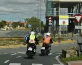 Dos motoristas con chalecos de prácticas, uno amarillo y otro naranja, circulan en moto. Uno de ellos lleva un cartel de "carnet moto 300".