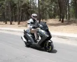 Hombre en casco y chaqueta conduce un triciclo de motor gris por una carretera rodeada de pinos.