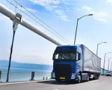 Un camión azul cruza un puente colgante, mostrando varios tipos de camiones en movimiento bajo un cielo azul.