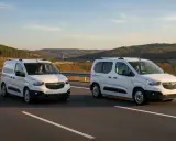 Dos furgonetas panel blancas circulan por una carretera con colinas boscosas al fondo.
