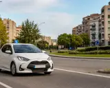 Coche blanco con señal "L" en el techo, listo para el traslado de expediente autoescuela. Edificios y árboles al fondo.