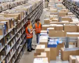 Tres trabajadores revisan inventario en un almacén, optimizando la logística de aprovisionamiento. Estanterías llenas de cajas y productos se extienden hasta el fondo.