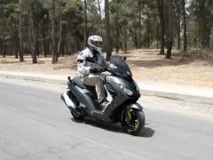 Hombre en casco y chaqueta conduce un triciclo de motor gris por una carretera rodeada de pinos.