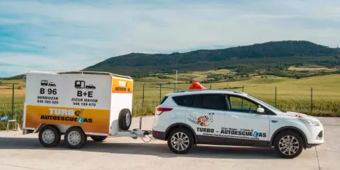 Un coche blanco de **4x4 autoescuela** con remolque, listo para enseñar a conducir en un paisaje rural.