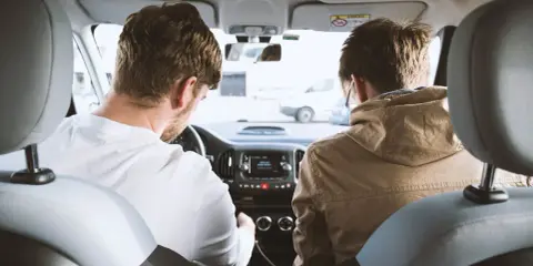 Dos hombres en un coche, uno conduce y el otro mira. Podrían estar discutiendo los tipos de faltas que se cometen al volante.