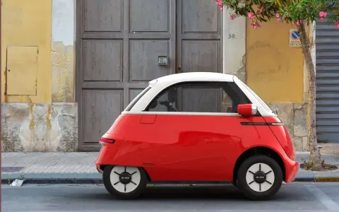 Un triciclo y cuadriciclo de motor rojo y blanco, un vehículo eléctrico compacto, estacionado frente a una puerta de madera y una pared amarilla.