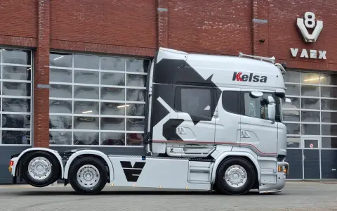 Camión Scania Longline plateado con diseño negro y rojo, aparcado frente a un edificio de ladrillo con puertas de garaje.