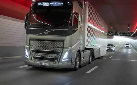 Camión Volvo FH Globetrotter plateado avanza por un túnel con luces rojas. El camión, que parece ser un modelo i-Save, transporta una carga con un diseño hexagonal.