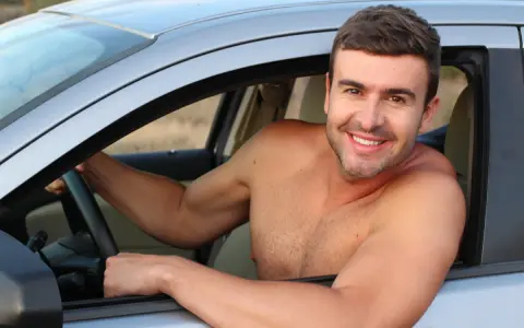 Hombre sonriente al volante de un coche, disfrutando de la libertad de que se puede conducir sin camiseta en un día caluroso.