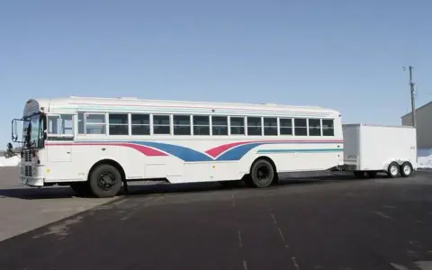 Autobús blanco con franjas azules y rosas, remolcando un tráiler. Ideal para un viaje largo con el carnet D+E.