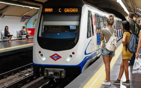 En la estación de metro, un tren blanco espera. Varias personas observan, una podría recibir una multa por usar tarjeta de transporte de otra persona.