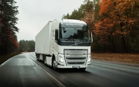Un camión Volvo FH blanco con remolque blanco circula por una carretera mojada rodeada de árboles otoñales.