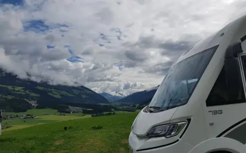Autocaravana de más de 3500 kg aparcada en un prado verde con vistas a un valle alpino y nubes dramáticas.