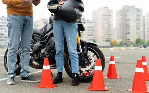Persona se prepara para el circuito examen moto a1. Un instructor revisa una lista junto a una moto y conos naranjas.