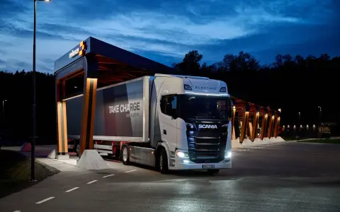Camión Scania eléctrico en una estación de carga nocturna. El remolque dice "TAKE CHARGE 100% ELECTRIC".